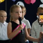 Dziewczynka trzyma telefon i wykonuje piosenkę razem z chorą koleżanką, uczestniczącą w uroczystości dzięki aplikacji messenger