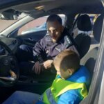 Chłopiec siedzi za kierownicą radiowozu, obok funkcjonariusz pokazujący wyposażenie samochodu