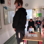 Chłopiec stoi tyłem do klasy, ma na uszacg słuchawki, próbuje usłyszeć co mówią do niego pozostali uczniowie