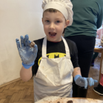 Chłopiec ubrany w czapkę kucharza i fartuch pokazuje wykonaną przez siebie pralinę czekoladową