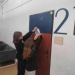 Dwie dziewczynki wieszają na drzwiach plakietkę z symbolem osób z niepełnopsrawnością