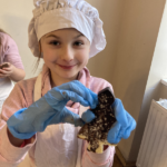 Dziewczynka w czapce kucharza i fartuszku z uśmiechem pokazuje do zdjęcia zrobioną przez siebie choinkę czekoladową