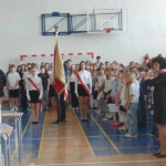 Uczniowie stoją na baczność i śpiewają hymn Polski, obok Pani gra na flecie