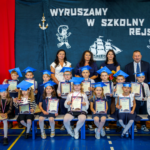 Dzieci w biretach i z medalami siedzą w dwóch rzędach, za nimi Pani Wizytator, Pani Dyrektor, Pan Dyrektor oraz Wychowawczyni