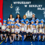 Dzieci w biretach z medalami i dyplomami siedzą w dwóch rzędach, za nimi Pani Wizytator, Pani Dyrektor, Pan Dyrektor oraz Wychowawczyni