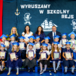 Dzieci w założonych biretach i z medalami siedzą w dwóch rzędach, za nimi Pani Wizytator, Pani Dyrektor, Pan Dyrektor oraz Wychowawczyni