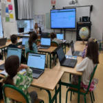 Dzieci siedzą przy stolikach i pracują na laptopach. W tle tablica multimedialna