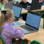 Dzieci siedzą przy stolikach i wykonują zadania na laptopach