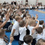 Dzieci siedzą na sali gimnastycznej i się zgłaszają