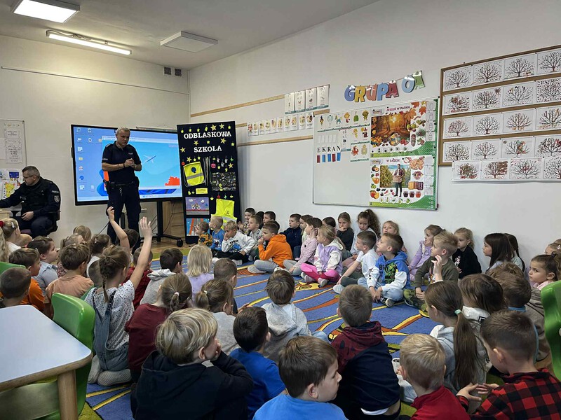 Dzieci z oddziałów przedszkolnych siedzą w kole słuchając dobrych rad pana policjanta. Widać dzieci zgłaszające się do zadania pytania. W tle odblaskowa gazetka.