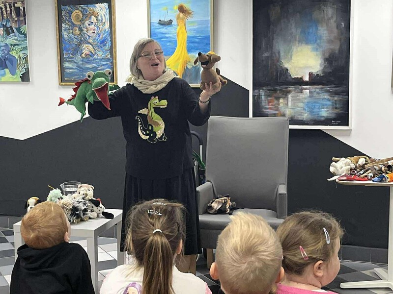 Ewa Stadtmüller – znana autorka książek dla dzieci, opowiada przedszkolakom ciekawe historie pokazując swoje kukiełki. Na ścianie w tle widać różne obrazy.