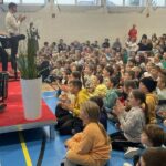 grupa dzieci siedzi przed sceną na której stoi meżczyzna