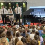 dwóch mężczyzn stoi na scenie, pośrodku stoi chłopiec, który będzie grał na instrumencie