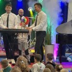 dwóch mężczyzn stoi na scenie a pośrodku nich stoi chłopiec, który ma ubraną kolorową perukę i gra na instrumencie