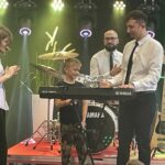 mężczyzna i chłopiec stoją na scenie za instumentem, z tyłu siedzi mężczyzna, z lewej strony stoi dziewczyna która klaszcze