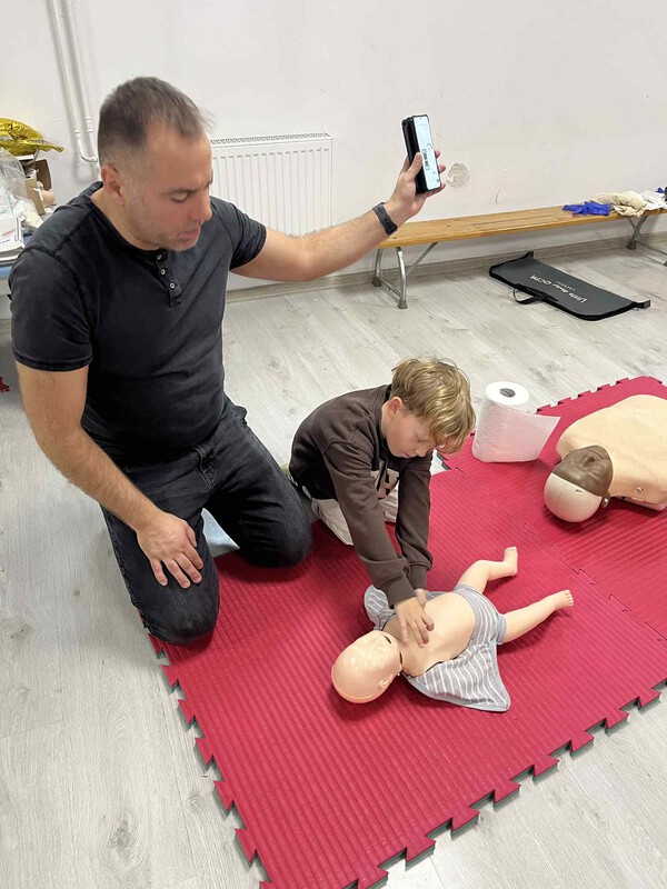 Przedszkolaki pod okiem ratownika ćwiczą masaż serca na fantomie niemowlaka.