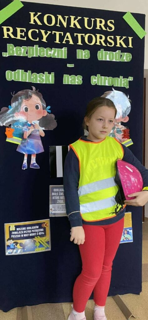 Dziewczynka ubrana w kamizelkę odblaskową i z kaskiem w ręku recytuje wiersz