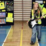 Dziewczynka ubrana w kamizelkę, trzyma gitarę prezentuje strój odblaskowy.