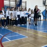 Dwoje uczniów siedzi i gra na instrumentach, obok oraz z tyłu stoją uczniowie, którzy śpiewają piosenki, przed nimi stoi pani dyrektor, która przemawia.