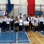 Dwóch uczniów siedzi na krzesłach i gra na instrumentach, obok oraz z tyłu stoją uczniowie którzy śpiewają.