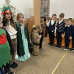 Od lewej strony stoi chłopiec i dziewczynka przebrani za elfy, obok stoi dziewczynka w stroju wróżki, na podłodze klęczą dwie dziewczynki przebrane za koty.Po prawej stronie stoi czterech chłopców z podniesionymi dłońmi do pasowania na czytelnika.