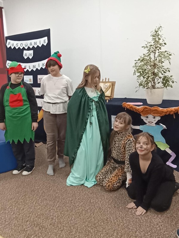 Po lewej stronie stoi chłopiec, obok stoi dziewczynka przebrani za elfy, obok stoi dziewczynka przebrana za wróżkę, obok klęczą dwie dziewczynki przebrane za koty.