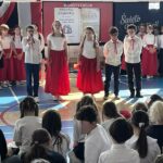 Dzieci na sali gimnastycznej stoją na tle dekoracji w trakcie apelu z okazji świąt majowych.